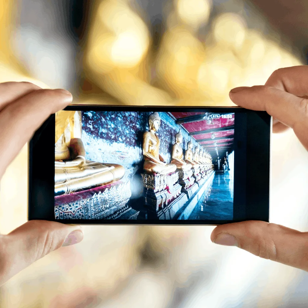 Two hands hold a smartphone, capturing a row of golden Buddha statues in a temple. The image conveys a sense of tranquility and cultural reverence.