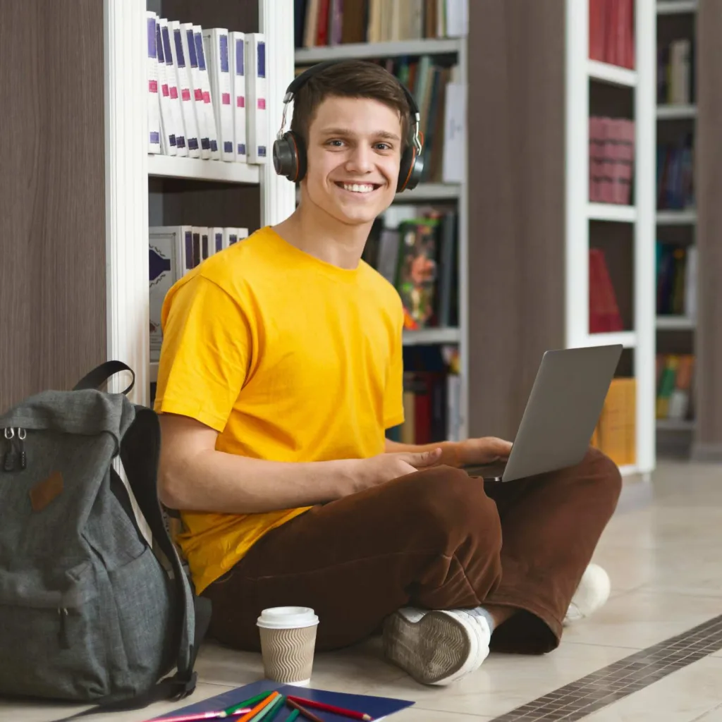 A young man in a yellow shirt sits on the floor in a library, wearing headphones and using a laptop. A backpack, coffee cup, and scattered pencils are nearby.